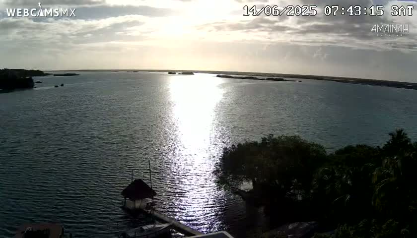 A tranquil body of water reflects the bright sunlight under a partly cloudy sky, with lush green vegetation visible along the shoreline and a small thatched structure on a dock.
