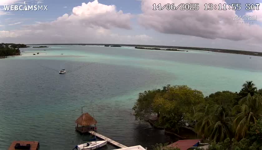 A partly cloudy day shows a tranquil body of teal water with small boats, a thatched roof structure on a dock, and lush green vegetation along the shoreline.