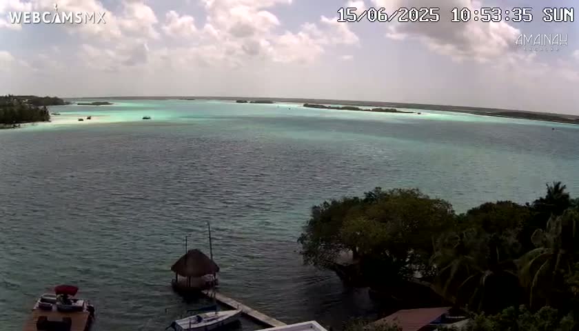 A partly cloudy sunny day overlooks a calm body of teal water with small islands and boats visible in the distance, and some boats and a small structure near the shoreline.