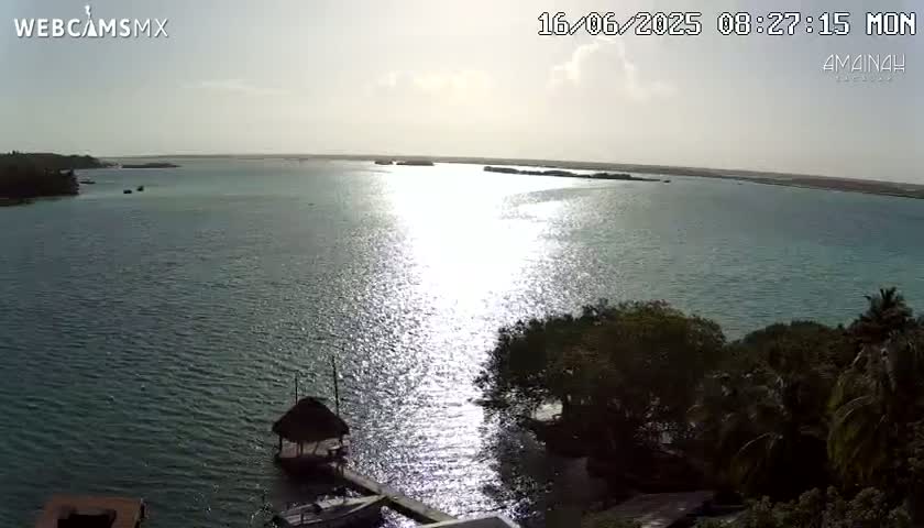 A tranquil body of water, sparkling under a bright, mostly sunny sky, is surrounded by lush green vegetation and features several small boats near a thatched-roof structure on the shore.