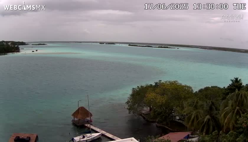 An overcast sky hangs over a calm body of teal water, where a small thatched-roof hut and sailboats are moored near a shoreline lined with lush green trees and palm trees.