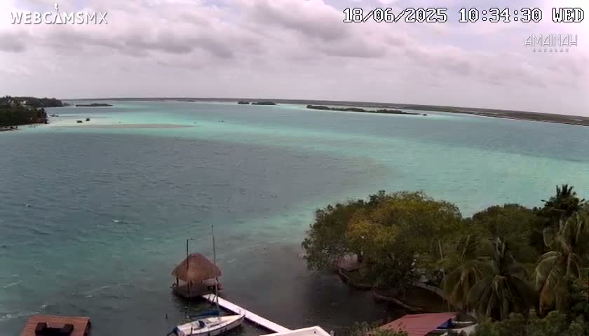 An expansive, teal-colored lagoon is partially shrouded by overcast skies, with lush green vegetation lining its shores and a small thatched-roof structure visible near a docked sailboat.