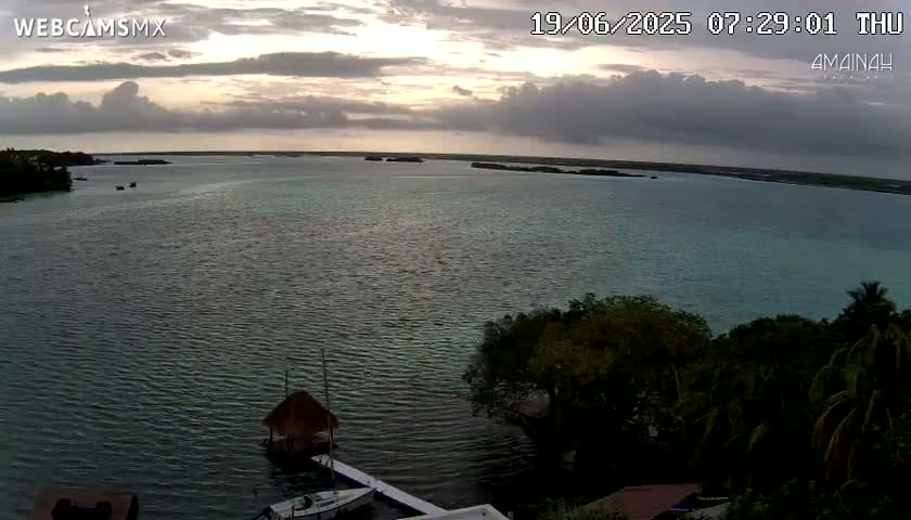 A calm body of water with a few boats and a thatched-roof structure is seen from an elevated vantage point under a partly cloudy sky at sunset.