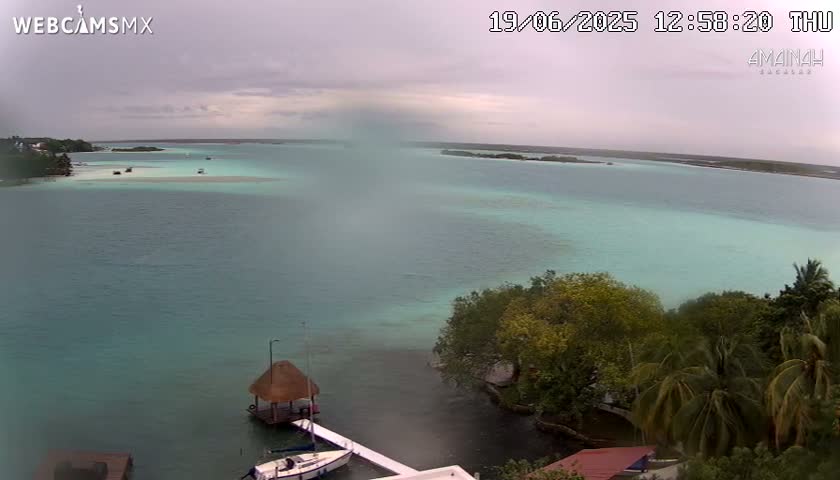 An overcast day reveals a tranquil turquoise lagoon, dotted with small boats and surrounded by lush green vegetation that descends to a waterfront with a thatched-roof gazebo and docked sailboats.