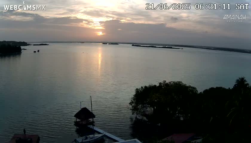 A calm body of water reflects a setting sun under a partly cloudy sky, with trees and small structures visible on the shore.