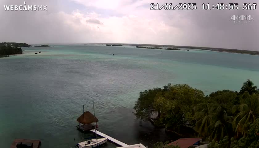 An overcast sky hangs over a calm, turquoise body of water with small islands in the distance, a few boats near the shore, and lush green vegetation lining the water's edge.