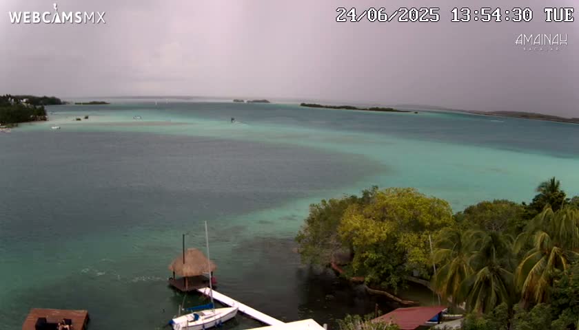 An overcast sky hangs over a calm body of teal water, where small boats are moored near a shoreline covered in lush green vegetation.