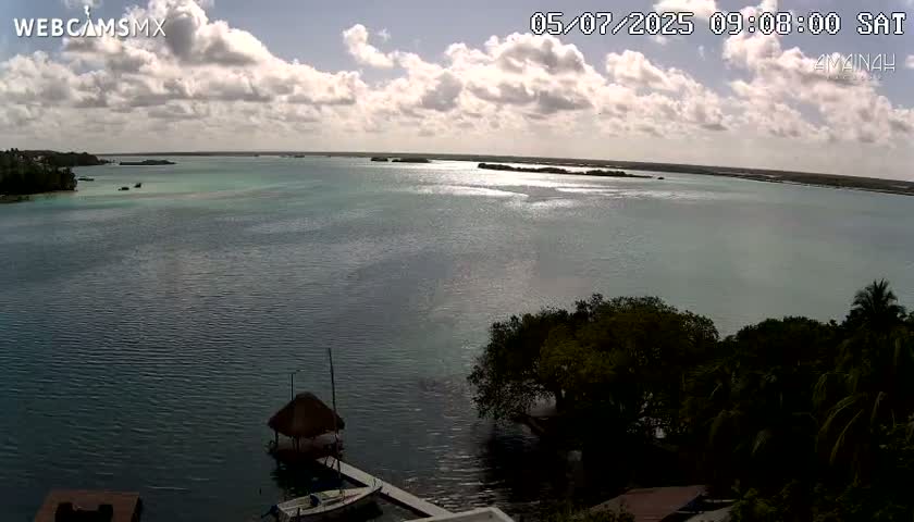 A partly cloudy sky overlooks a calm body of water with small islands in the distance, and tropical vegetation in the foreground near a dock with a few small boats.