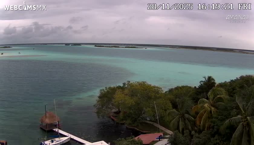 A wide view of a large, multi-hued blue lagoon with distant landmasses, a dock with a sailboat, and lush tropical vegetation in the foreground, all under a cloudy, overcast sky.