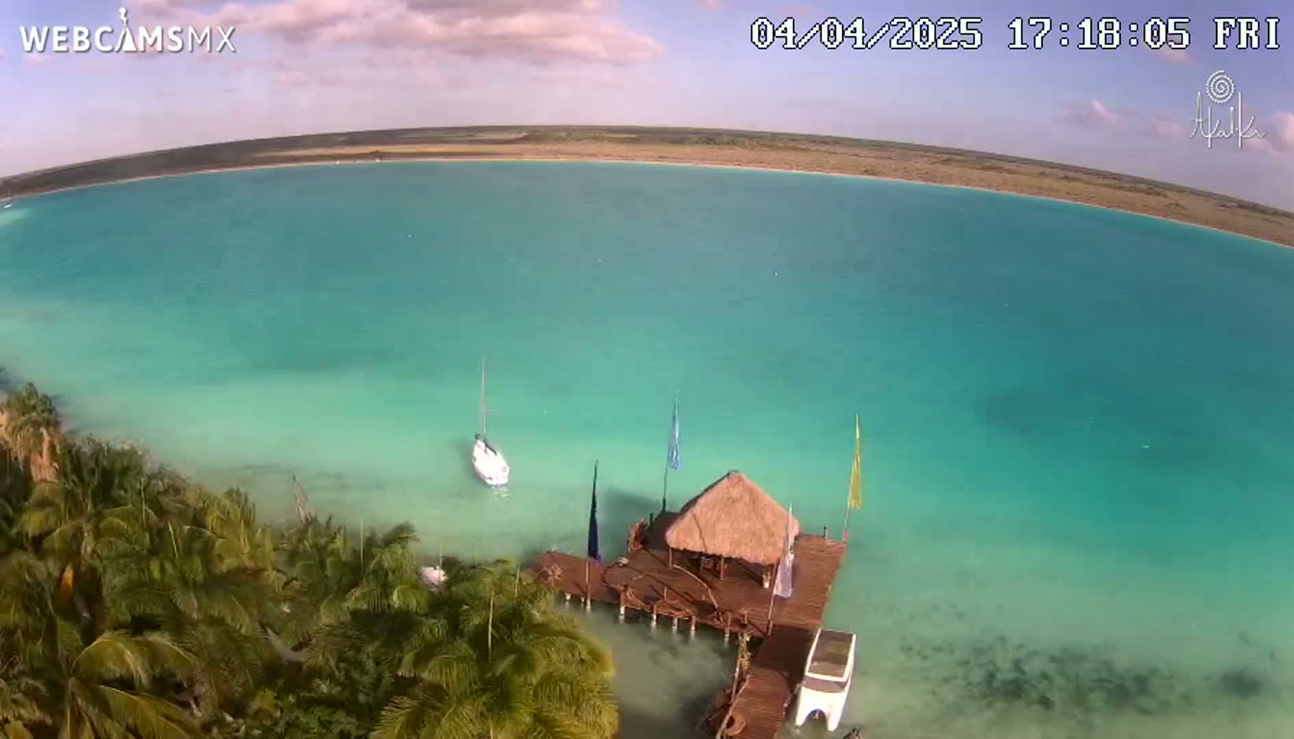An aerial view shows a calm, bright turquoise body of water with a small sailboat and a thatched-roof hut on a dock, surrounded by palm trees under a partly cloudy sky.