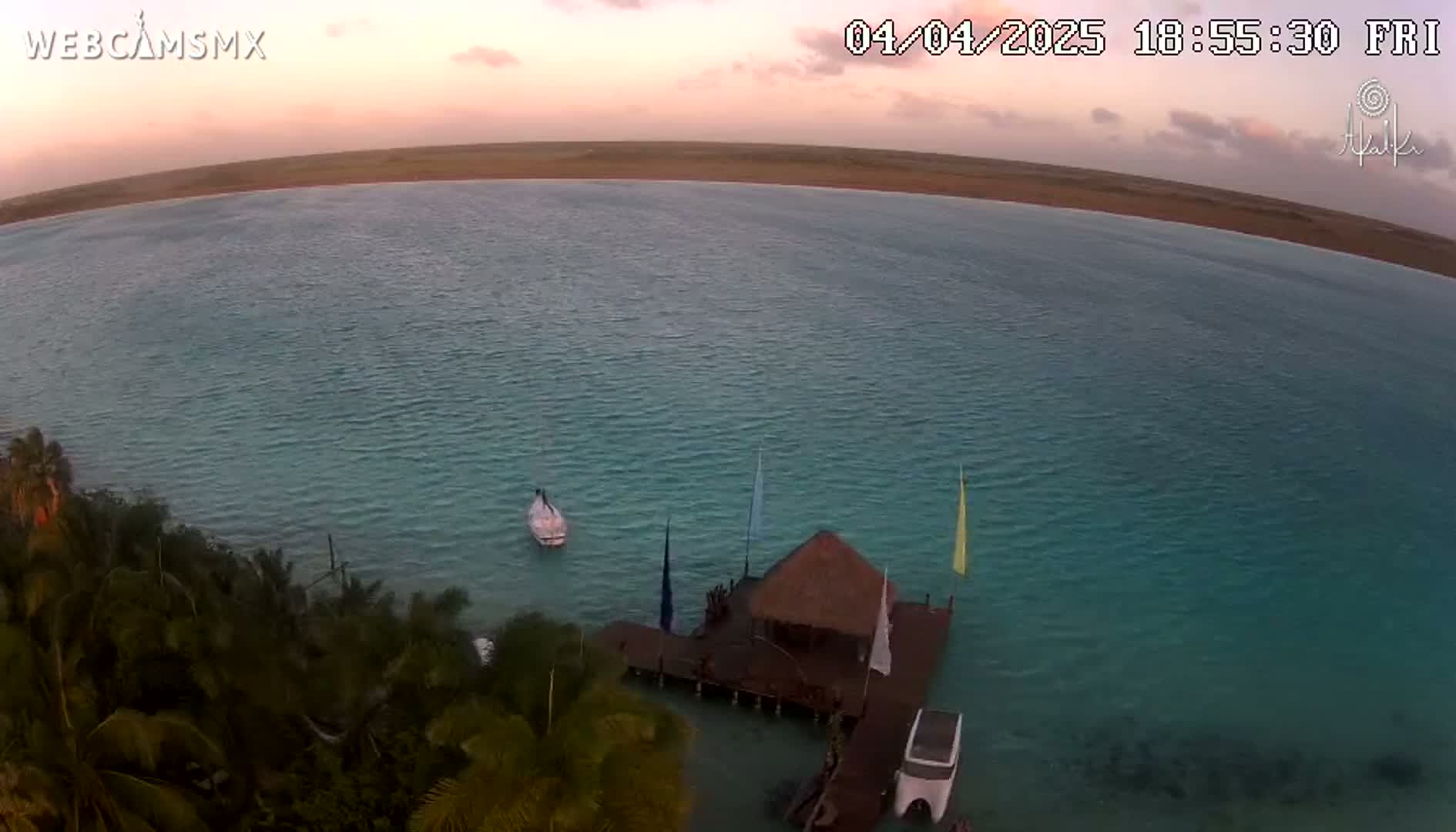 An aerial view shows a calm, teal body of water at sunset with a thatched-roof structure on a dock, a small boat, and lush tropical vegetation along the shore.