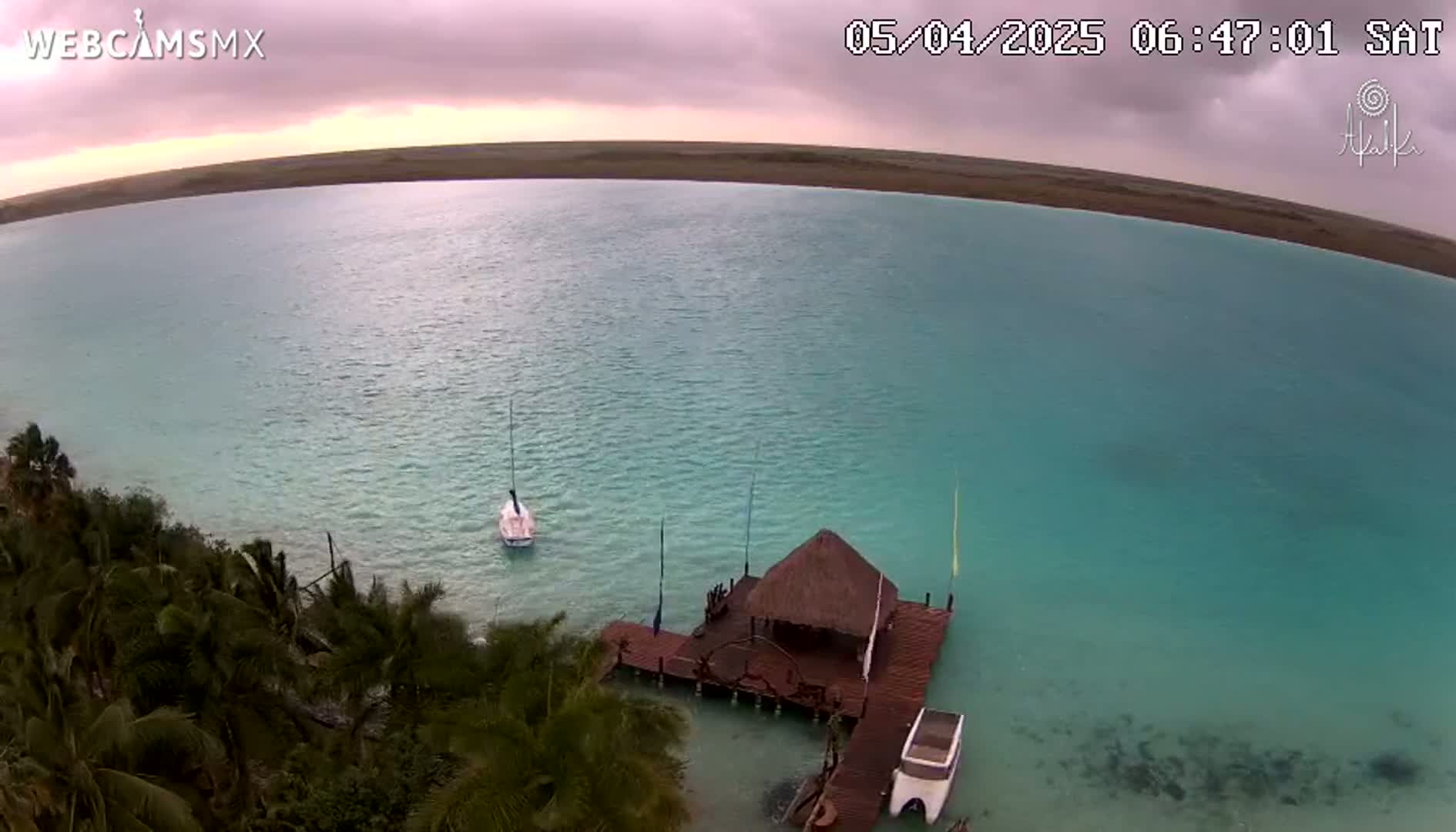 An aerial view shows a calm, turquoise body of water with a small sailboat and a thatched-roof structure on a dock, surrounded by lush green vegetation under an overcast sky.