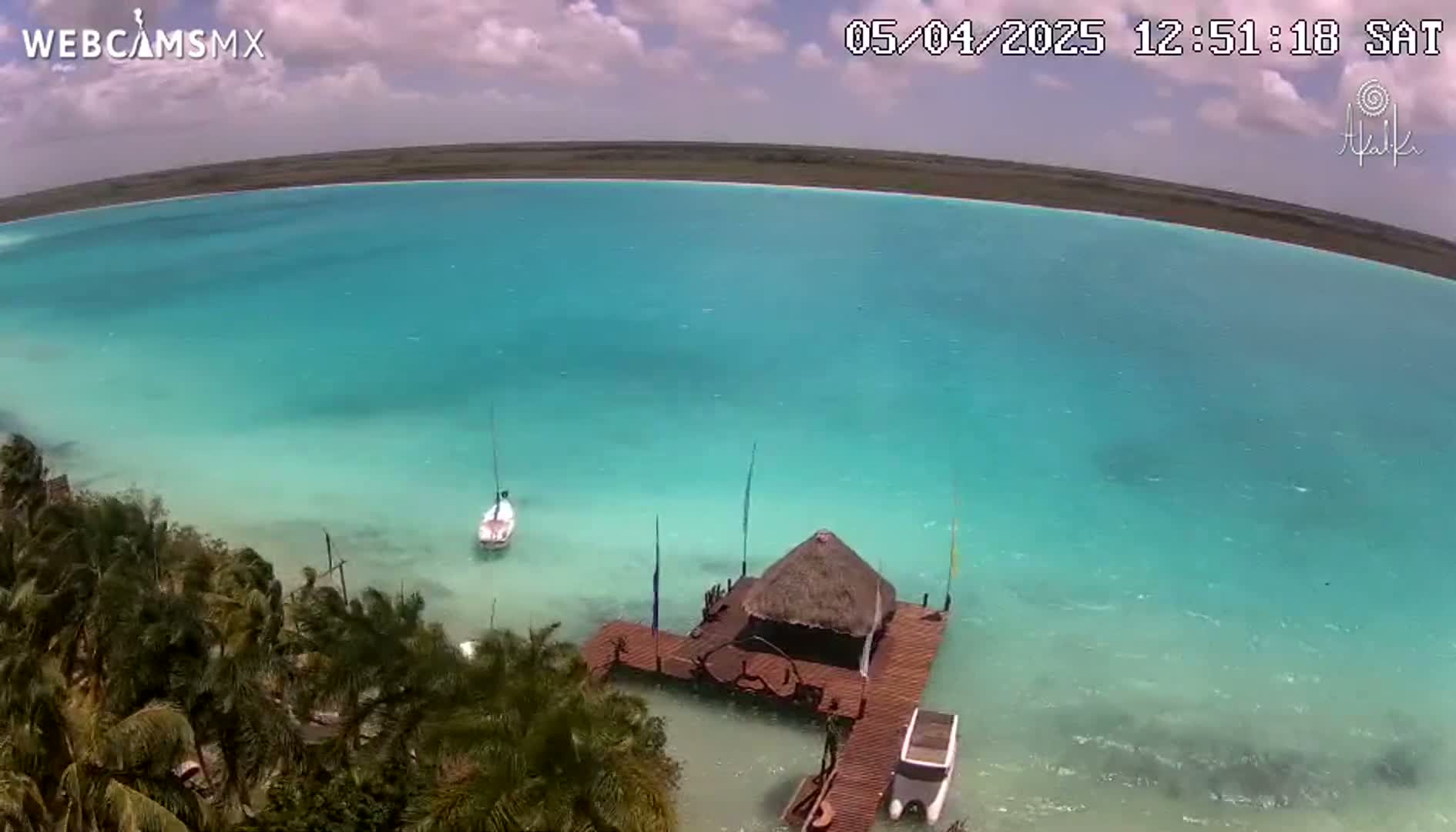 An aerial view shows a bright turquoise body of water with a small sailboat and a thatched-roof hut on a dock, under partly cloudy skies.