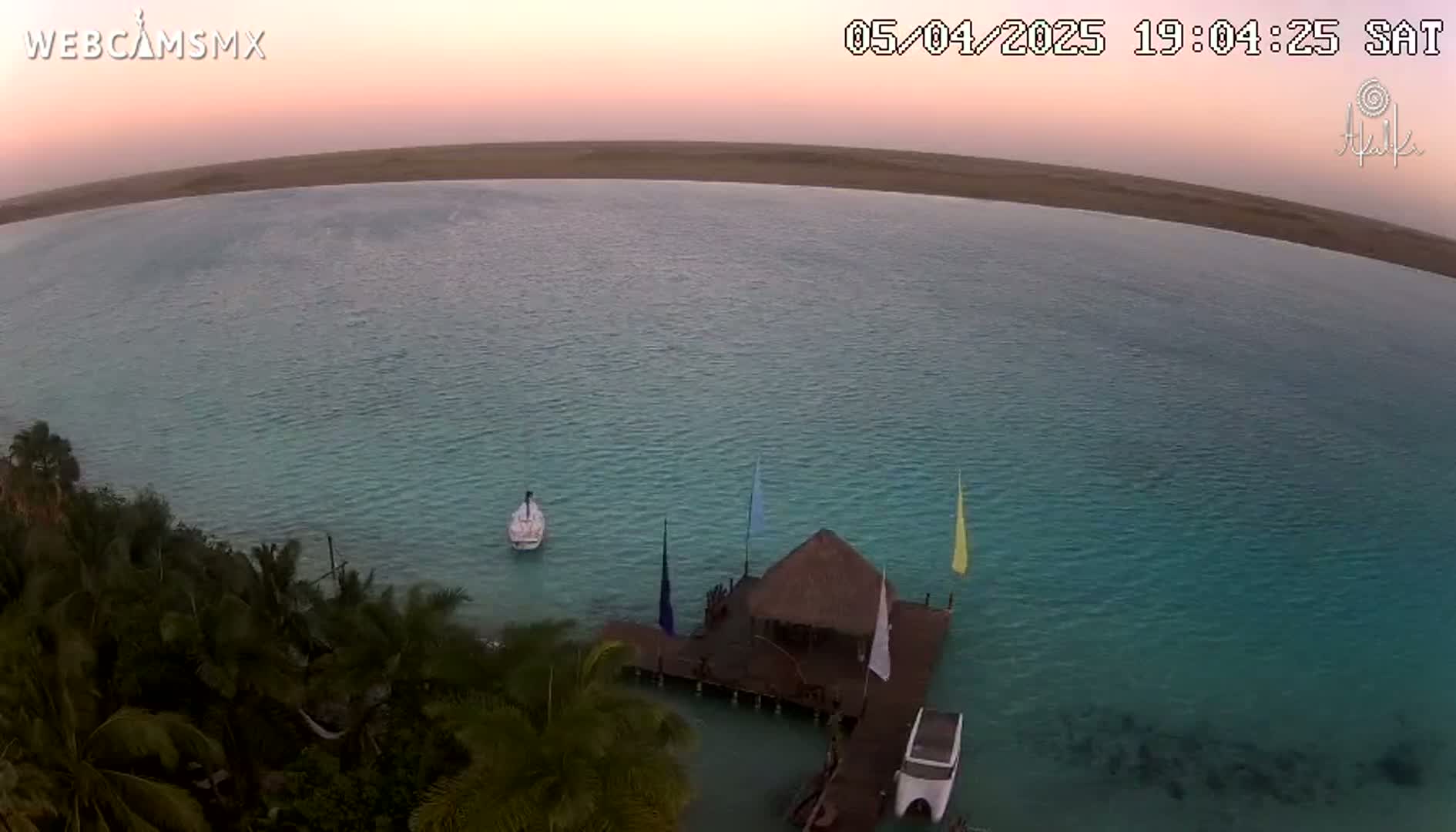 An aerial view shows a calm, turquoise body of water at sunset, with a small thatched-roof structure on a dock, a few boats, and lush tropical vegetation on the shore.