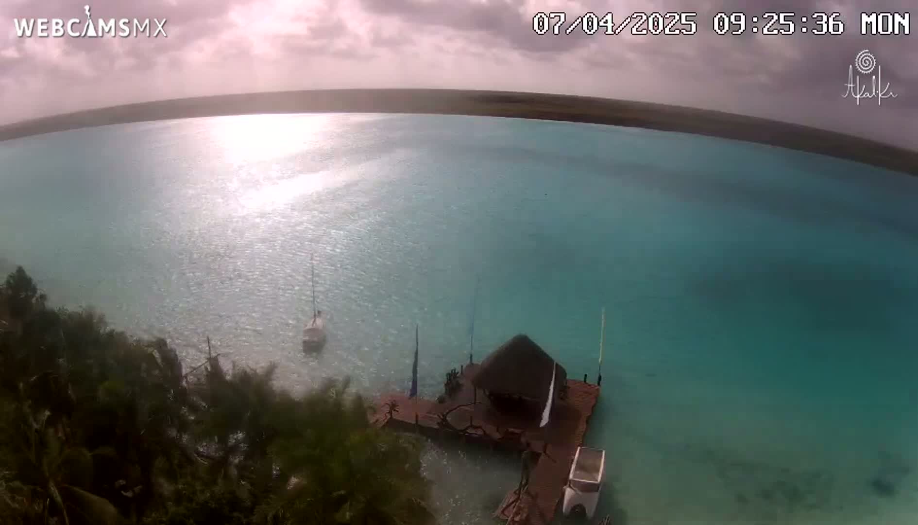 An aerial view shows a partly sunny day over a calm, turquoise body of water with a small dock and two sailboats, surrounded by lush green vegetation.