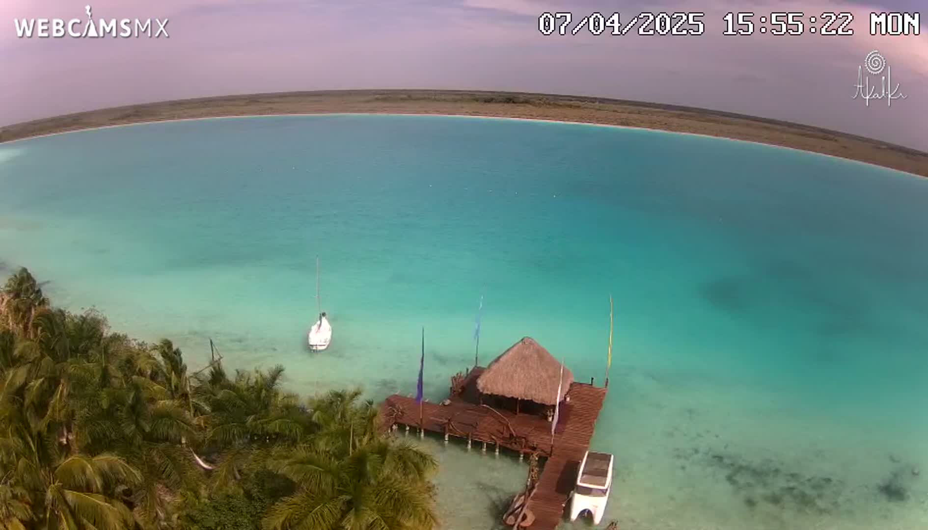 An aerial view shows a calm, turquoise body of water with a small sailboat and a thatched-roof hut on a wooden dock, surrounded by palm trees under a partly cloudy sky.