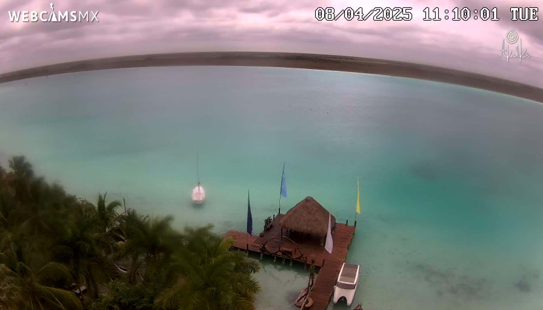 An elevated view shows a calm, teal-colored body of water under an overcast sky, featuring a small sailboat, a thatched-roof structure on a dock, and lush tropical vegetation along the shoreline.