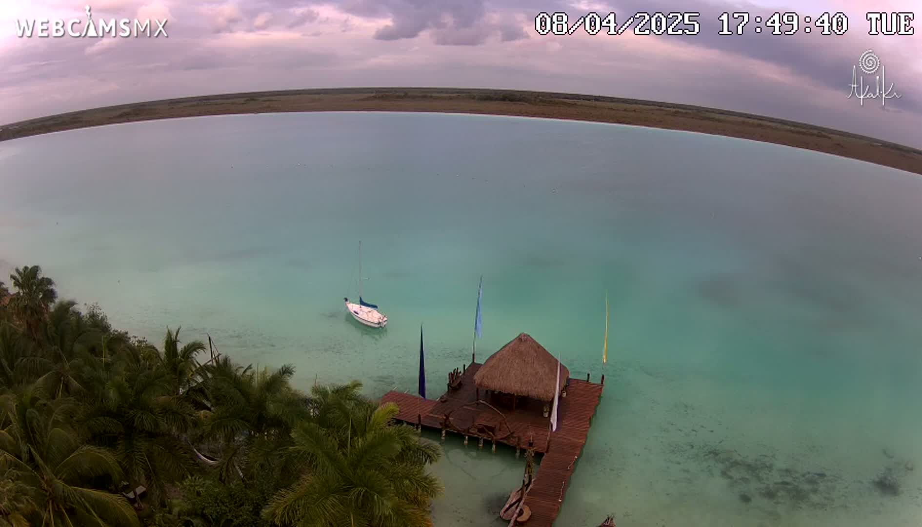 An aerial view shows a tranquil, light teal body of water under an overcast sky, with a small sailboat and a thatched-roof hut on a dock surrounded by palm trees.