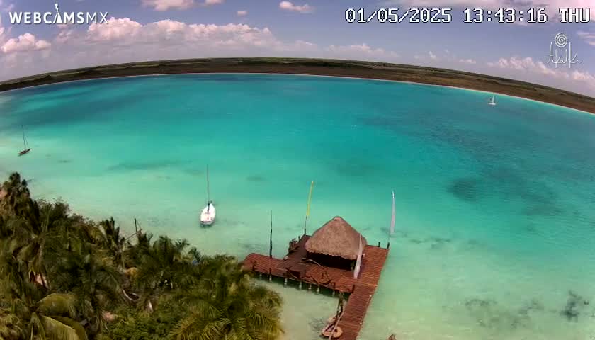 An aerial view shows a tranquil, bright turquoise body of water with a few sailboats, a thatched-roof structure on a wooden dock, and lush green vegetation along the shore under a partly cloudy sky.