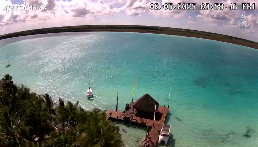 An aerial view shows a calm, turquoise body of water with several small boats near a wooden dock and thatched-roof structure, surrounded by lush green vegetation under a partly cloudy sky.