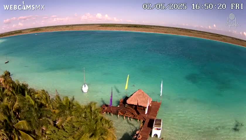 An aerial view shows a tranquil turquoise body of water with a thatched-roof structure on a dock, surrounded by lush palm trees under a partly cloudy sky.