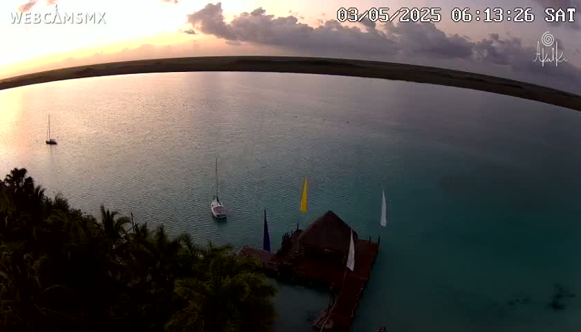 An aerial view shows calm, teal water with several sailboats near a thatched-roof structure on a dock at sunset under partly cloudy skies.