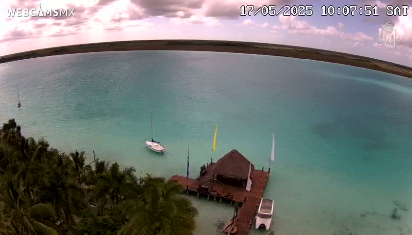 A tranquil turquoise lagoon is seen from an aerial perspective under partly cloudy skies, with a small sailboat and a thatched-roof structure on a dock visible near the shore, surrounded by lush palm trees.