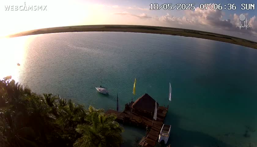 An aerial view shows a calm, teal-colored body of water at sunrise with a few sailboats, a small dock with a thatched-roof structure, and lush green vegetation along the shore.