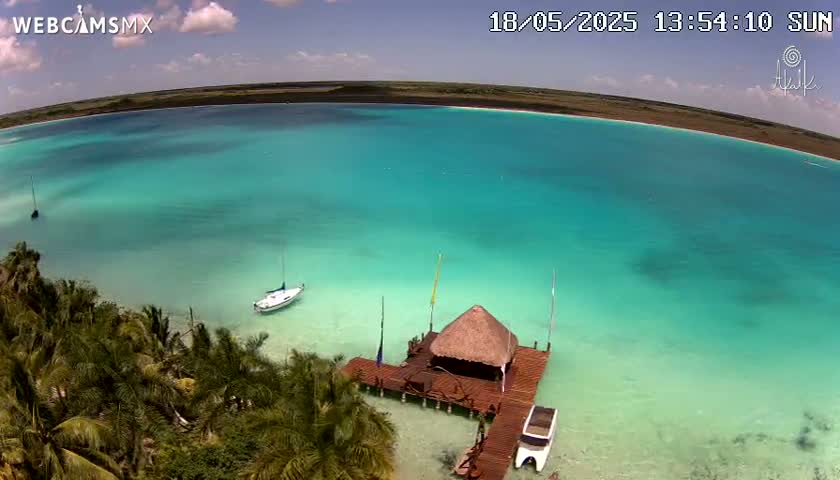 A sunny day shows a turquoise lagoon with a small sailboat and a thatched-roof hut on a wooden dock, surrounded by palm trees.