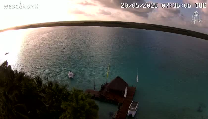 An aerial view shows a calm body of teal water under a partly cloudy sunrise sky, with a small thatched-roof structure on a dock and several small boats nearby.
