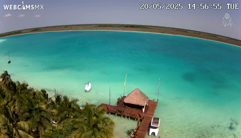 An aerial view shows a tranquil, turquoise lagoon on a sunny day, with a small thatched-roof structure on a wooden dock near a lush, green shoreline and several small boats.