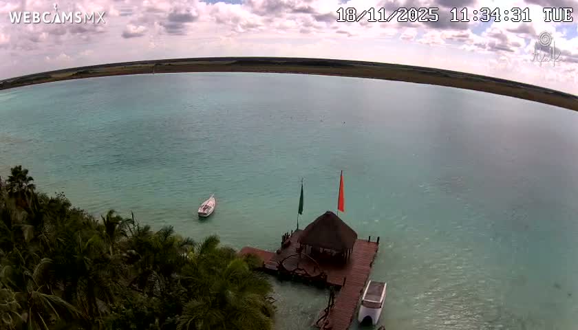 An aerial view shows a tranquil, turquoise lagoon on a sunny day, with a small thatched-roof structure on a wooden dock near a lush, green shoreline and several small boats.