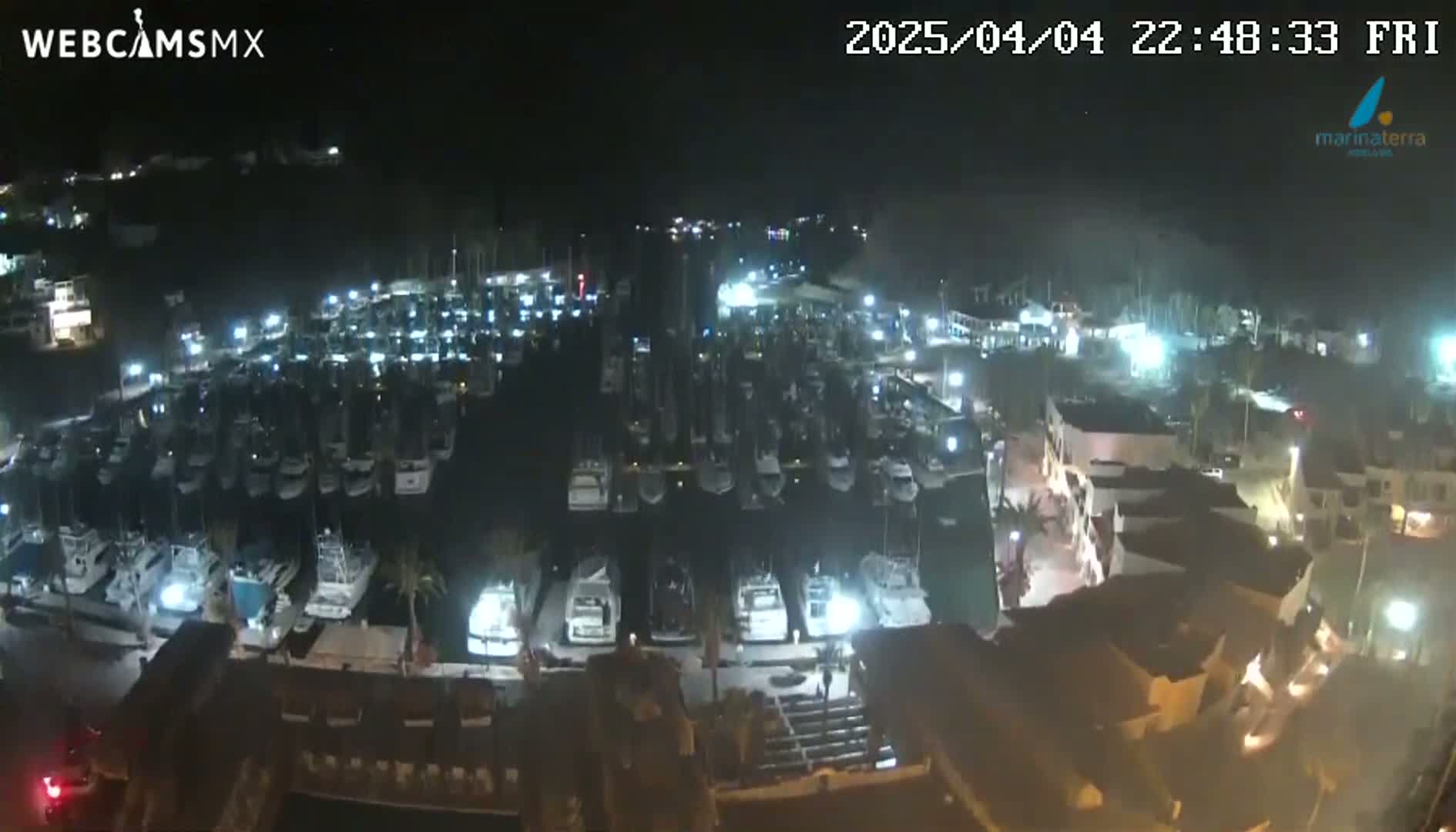 A nighttime aerial view shows a marina filled with many boats docked at the water's edge, next to buildings with lights on.