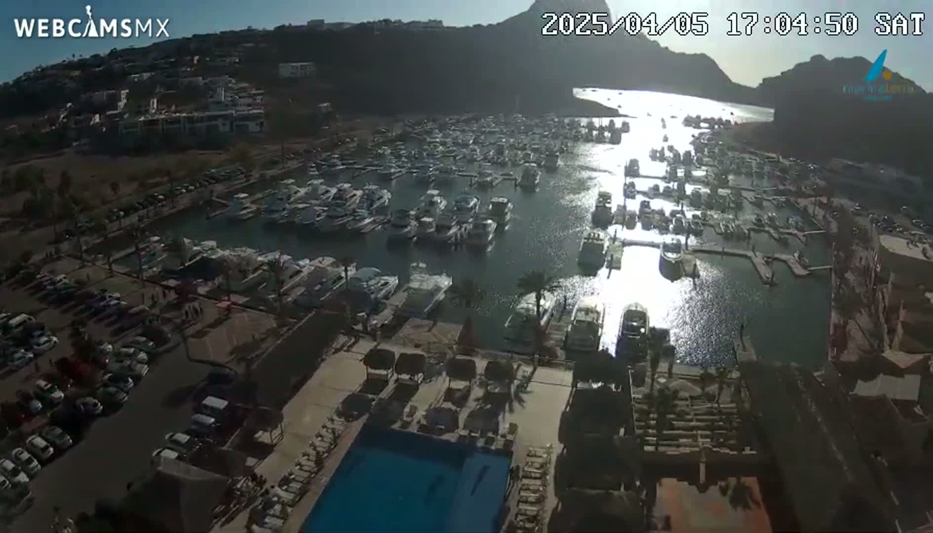 An aerial view shows a marina filled with boats, a parking lot, a resort pool, and surrounding buildings under a sunny sky.