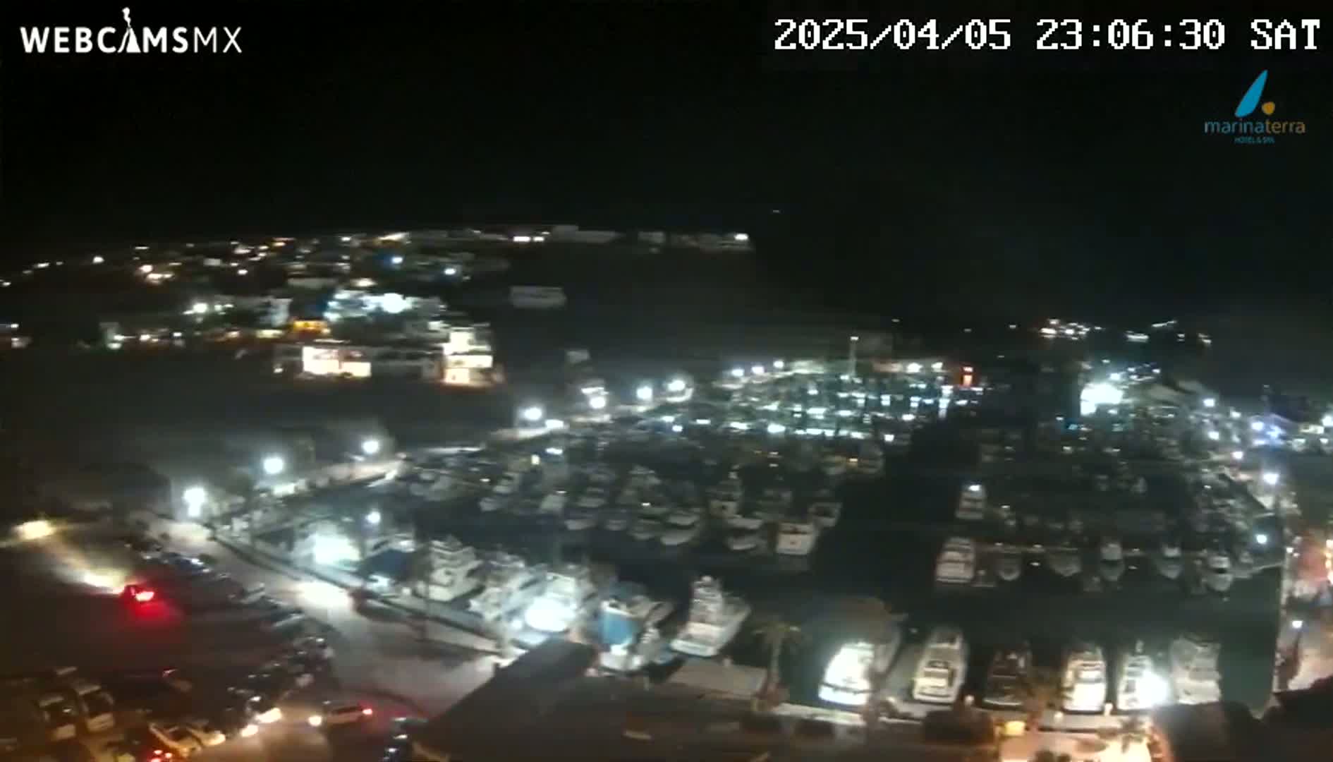A nighttime, high-angle view of a marina filled with numerous boats, surrounded by dimly lit buildings and roads with some cars parked.
