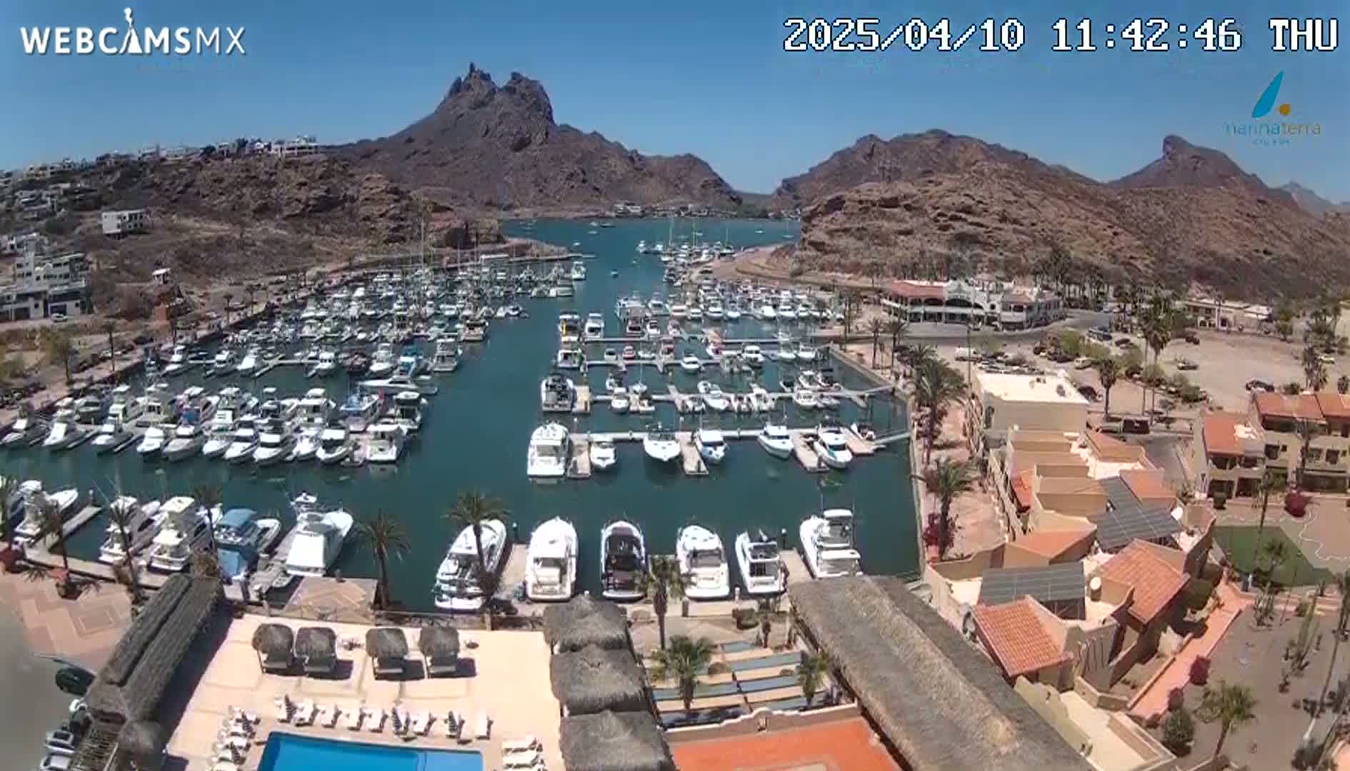 A marina filled with numerous boats is situated in a bay surrounded by arid, rocky hills under a clear, sunny sky.