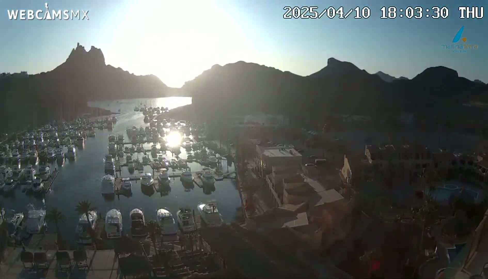 A marina filled with numerous boats is nestled between mountains, with the sun rising or setting, casting a golden light across the water.