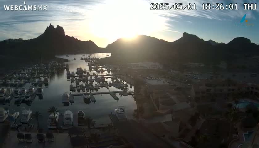 A sunlit harbor filled with numerous boats is nestled between mountains, under a partly cloudy sky.