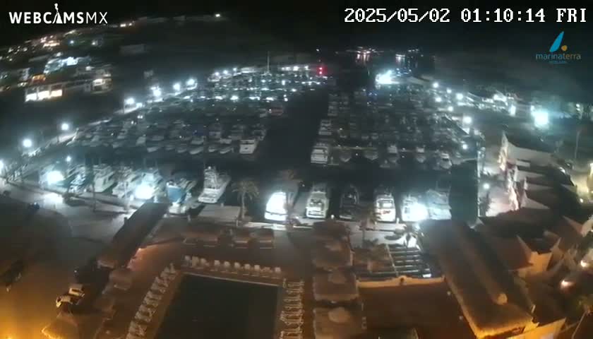 A nighttime aerial view of a marina filled with many boats, adjacent to buildings with outdoor furniture and a swimming pool.