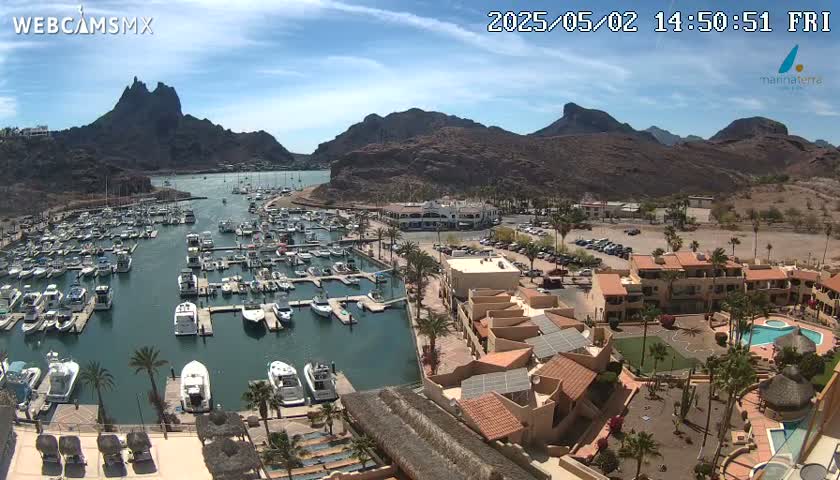 A marina filled with boats is situated next to a complex of low-rise buildings and a swimming pool, all set against a backdrop of arid, rocky mountains under a mostly sunny sky.
