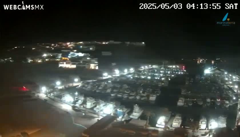 A nighttime aerial view shows a large marina filled with many boats, illuminated by lights, under a clear, dark sky.