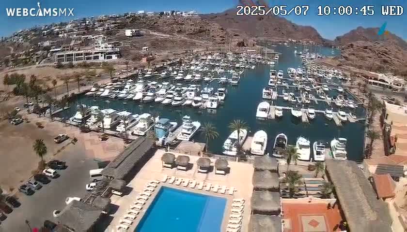 An aerial view shows a marina filled with boats on a sunny day, next to a resort with a swimming pool and lounge chairs.