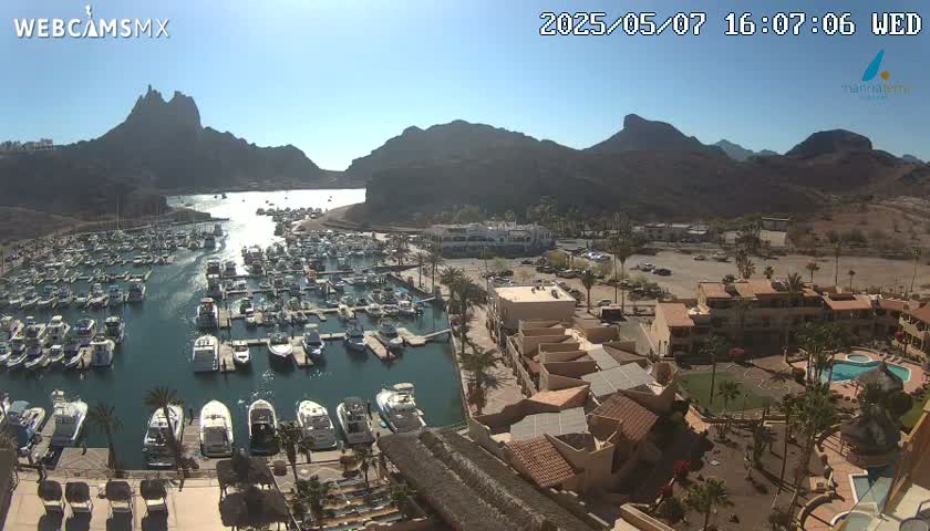 A sunny day overlooking a marina filled with numerous boats, nestled between arid, mountainous terrain and a resort complex with a swimming pool.