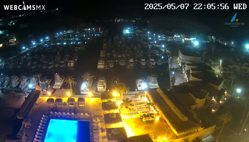A nighttime aerial view shows a marina filled with boats, a resort area with a lit pool and outdoor seating, and palm trees under a clear sky.
