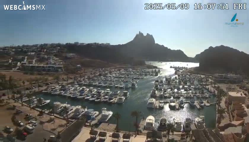 A sunny day shows a marina filled with many boats nestled between a hillside of buildings and a large rocky mountain.