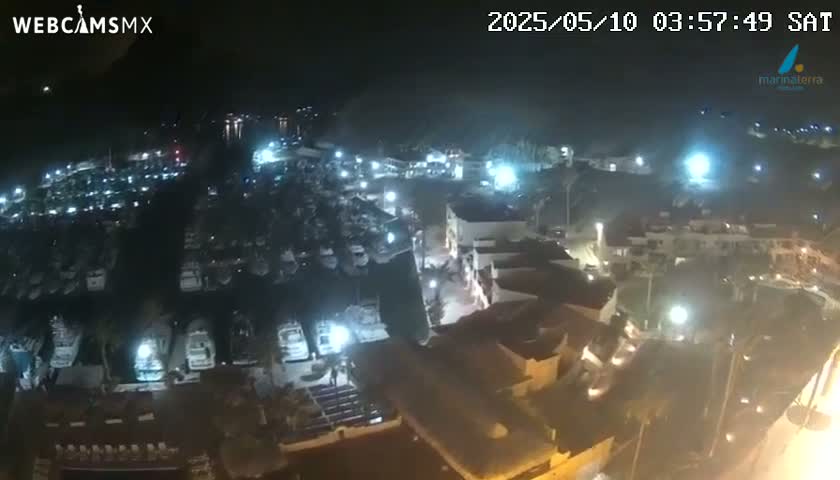 A nighttime aerial view shows a marina filled with boats and surrounding buildings illuminated by lights.