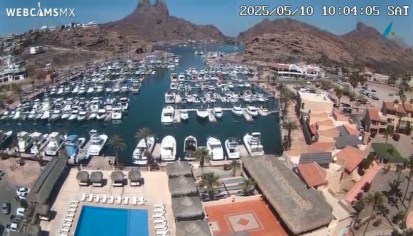 A marina filled with numerous boats is situated near a resort with a pool, all under a sunny sky and nestled against rocky mountains.