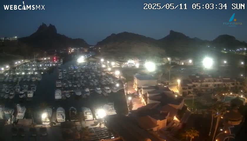 A nighttime aerial view shows a marina filled with boats, nestled between illuminated buildings and dark mountains under a twilight sky.
