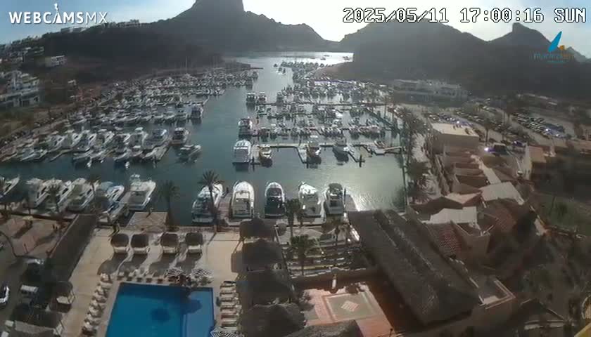 A sunny day overlooking a marina filled with boats, a resort pool, and buildings with terracotta roofs, nestled against a mountainous backdrop.