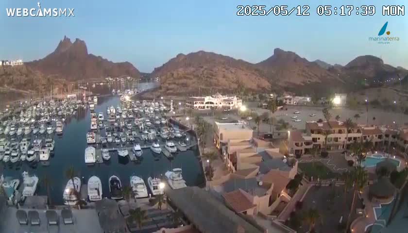 An aerial view shows a marina filled with boats at dusk, nestled between brown hills under a clear sky.